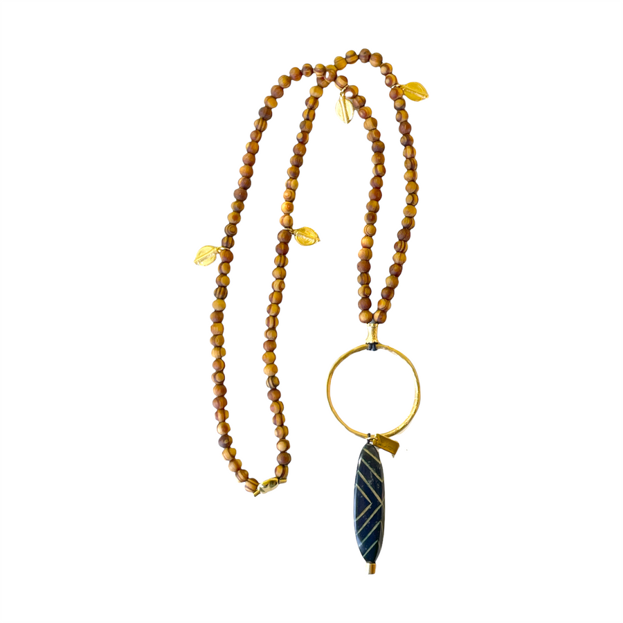 Collar largo de cuentas de madera y una pieza central compuesta de un aro de zamak con baño de oro mate y una espectacular pieza de madera pintada y lacada de inspiración africana. La pieza se complementa con unas cuentas de Zamak con baño de oro mate que dan vida a la madera.